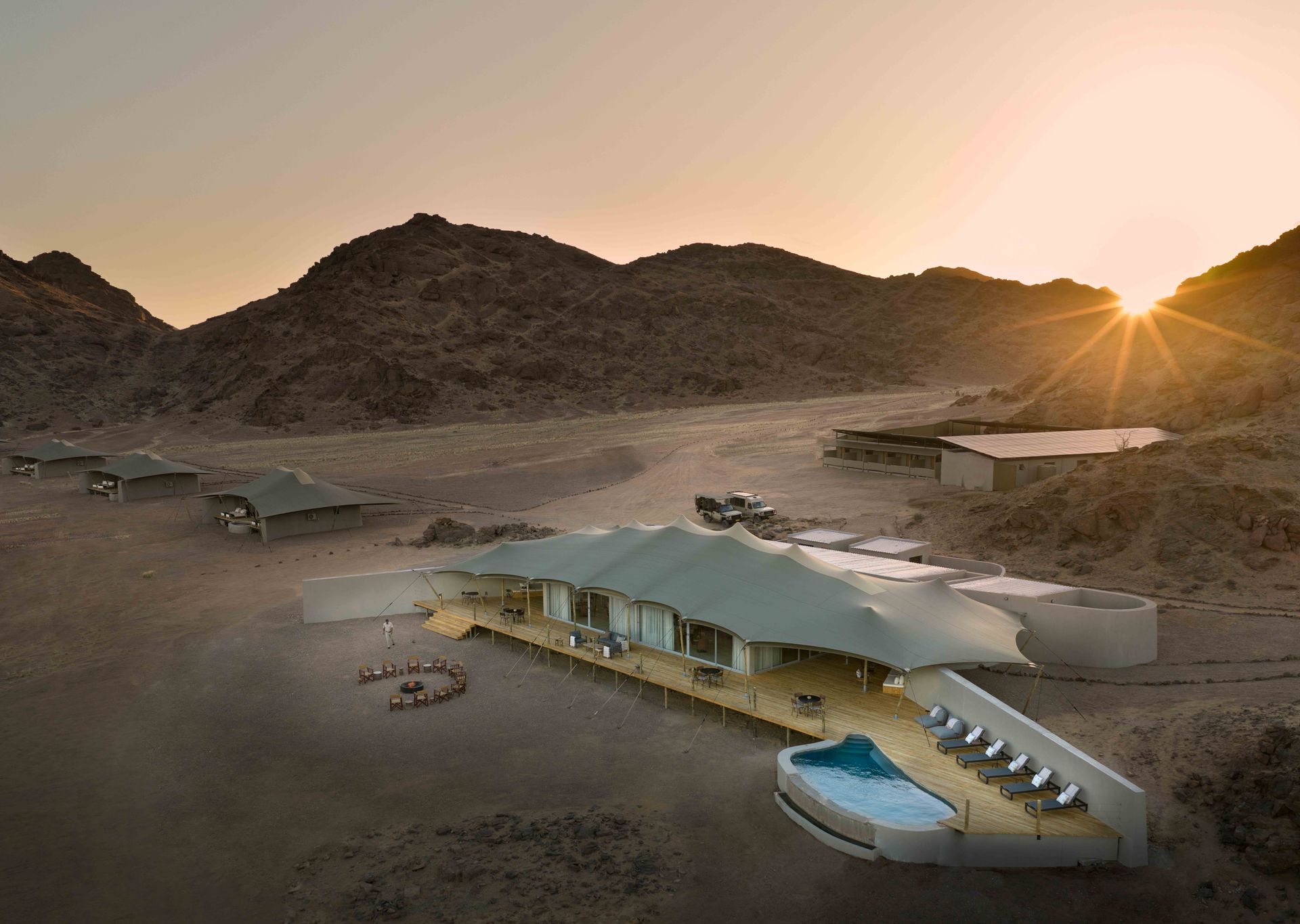 An aerial view of the Hoanib Skeleton Coast Camp at dusk with the hills on the horizon and the sun setting behind them